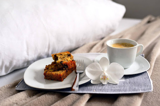 A full tea cup, a flower, and a slice of banana bread on a plate sitting on top of a bed