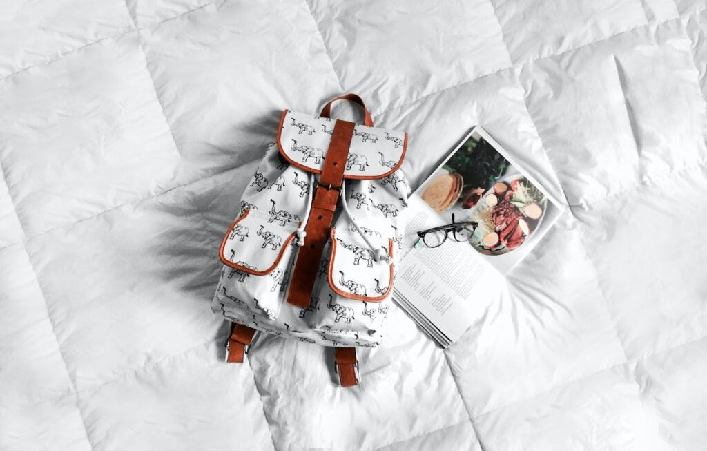 A white comforter with a small elephant print backpack, open book, and pair glasses on top