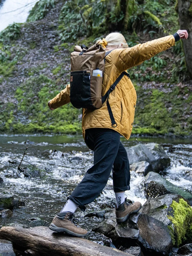 Davis Daypack in EPX Coyote color, perfect for everyday use, shown on a woman hiking over river rocks 