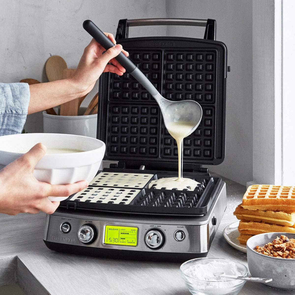 Non-Toxic Waffle Maker sitting open on a kitchen counter while a woman pours batter on top with a ladle