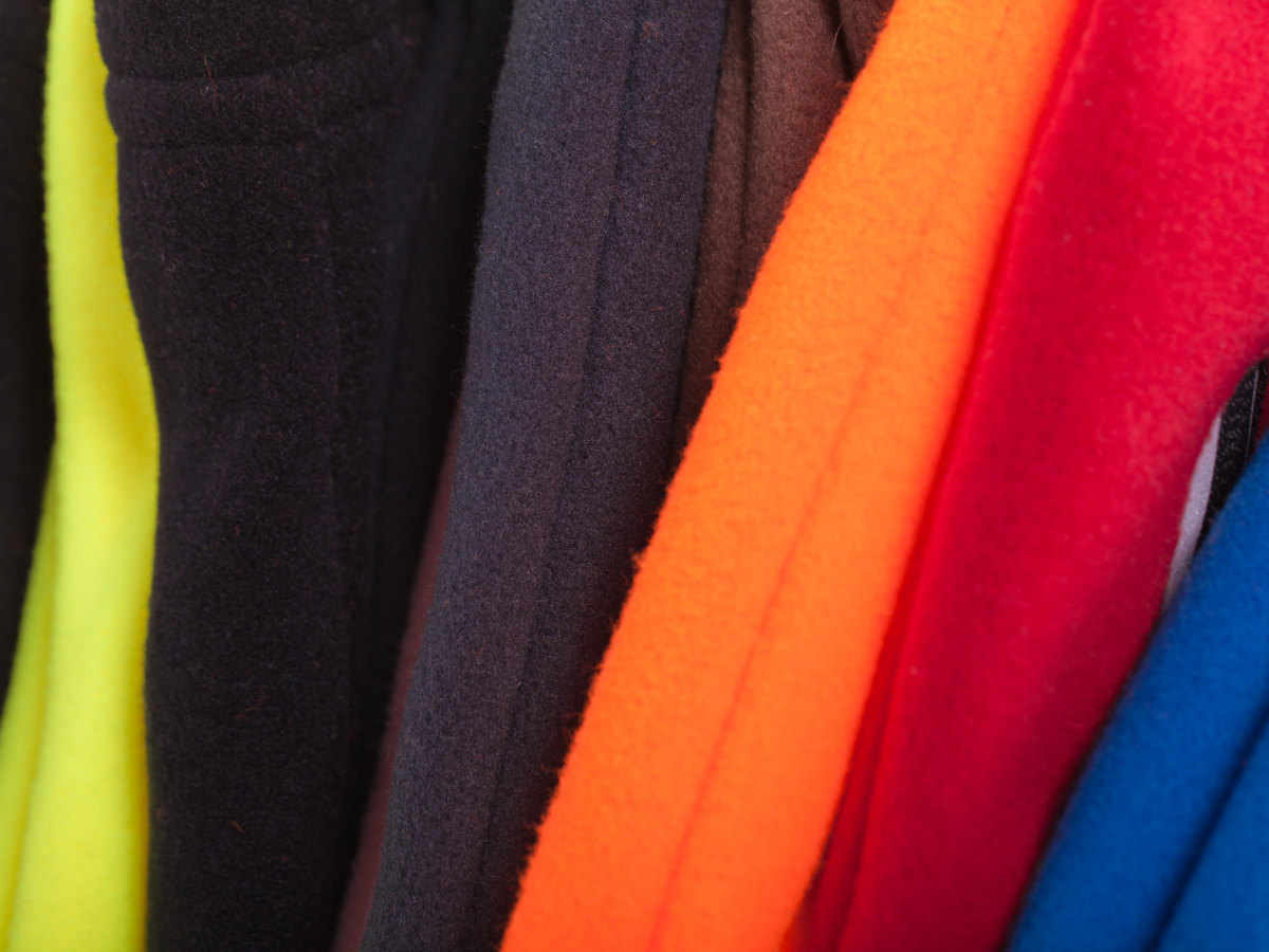 An array of fleece clothing, including pullovers and sweatpants, hang on a clothing rack. Colors are red, blue, black, yellow, and purple.