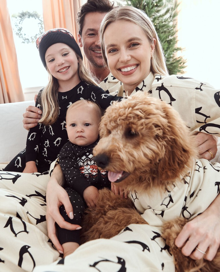 A family of four, including a mother, father, daughter, and son, as well as a family dog, sit on a white couch with matching penguin-themed pajamas.