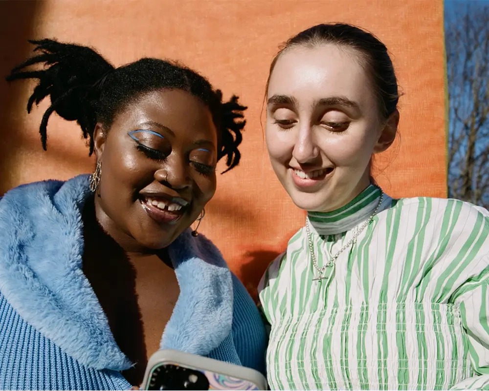 Two stylish women smiling while looking at a smartphone together, one wearing a blue fuzzy jacket and the other in a green striped top, with an orange backdrop and blue sky in the background.