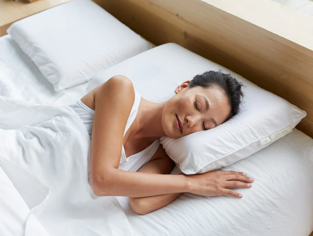 A woman lays on a bed with white sheets and pillows from Coyuchi
