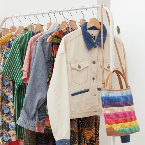 A clothing rack featuring a variety of colorful pre-loved garments, including floral patterns, striped dresses, denim, and a cream jacket with a blue collar, alongside a rainbow-striped woven bag.