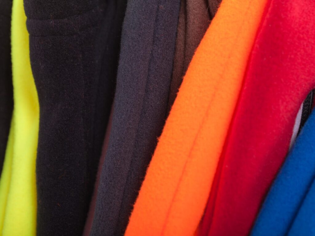 An array of fleece clothing, including pullovers and sweatpants, hang on a clothing rack. Colors are red, blue, black, yellow, and purple.