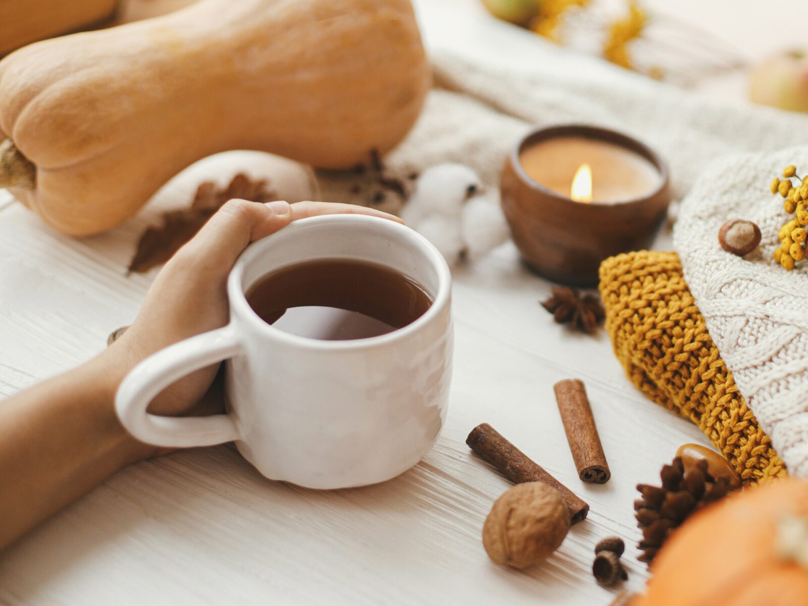 A hand holding a white mug full of coffee that’s sitting on a table with squashes, spices, knit sweaters, and a candle on top