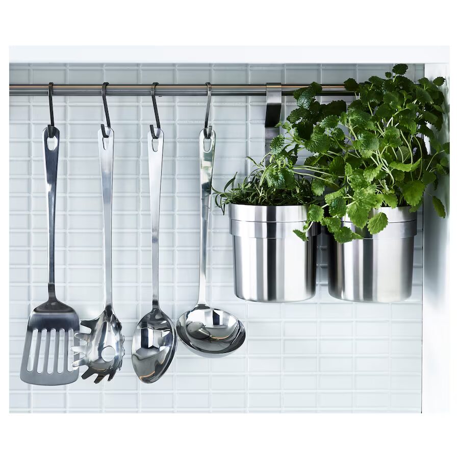 Non-toxic stainless steel kitchen utensils hanging on a rack beside fresh herbs in steel containers.