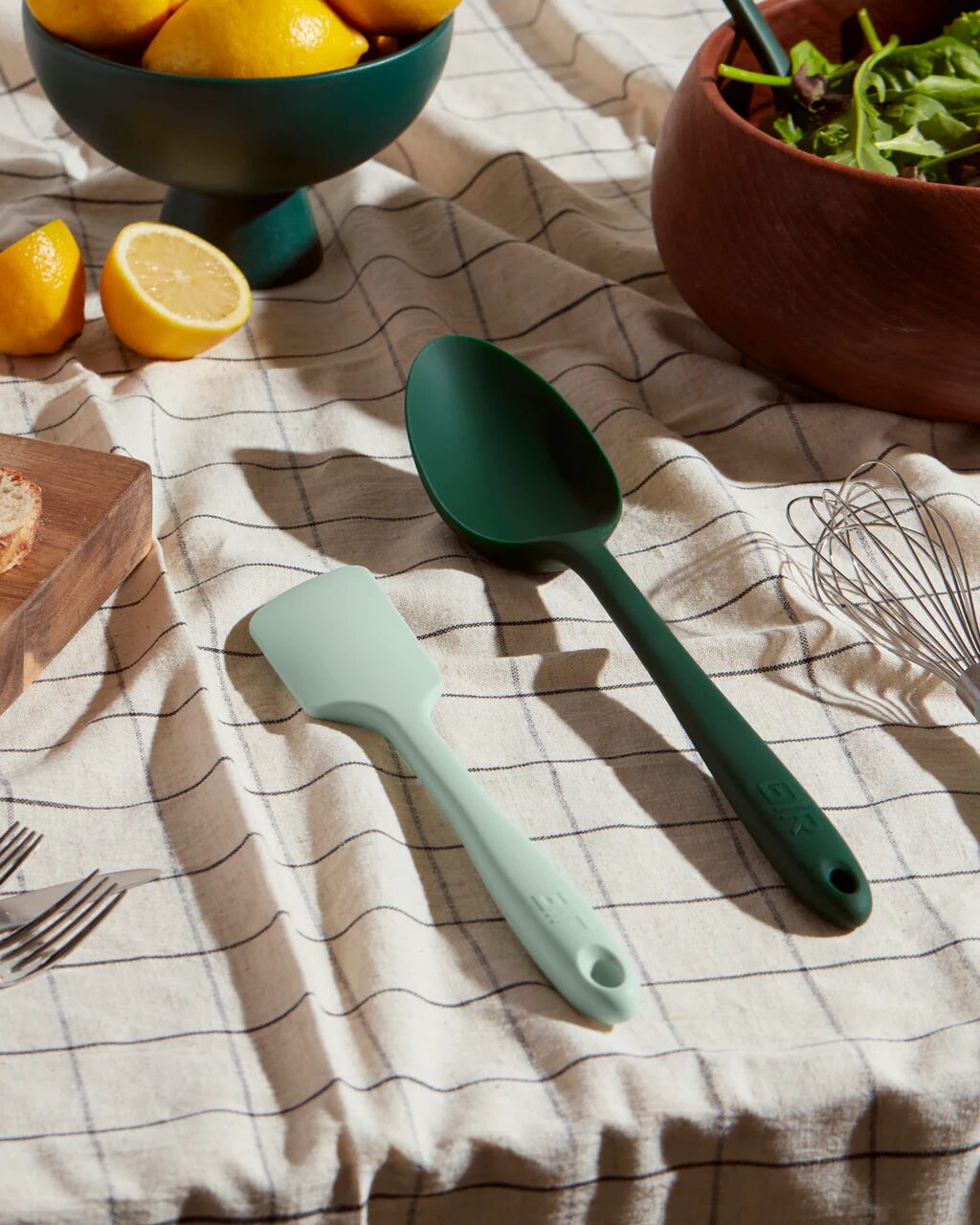 Non-toxic silicone kitchen utensils, including spatula and spoon, on a checkered cloth with fresh ingredients.