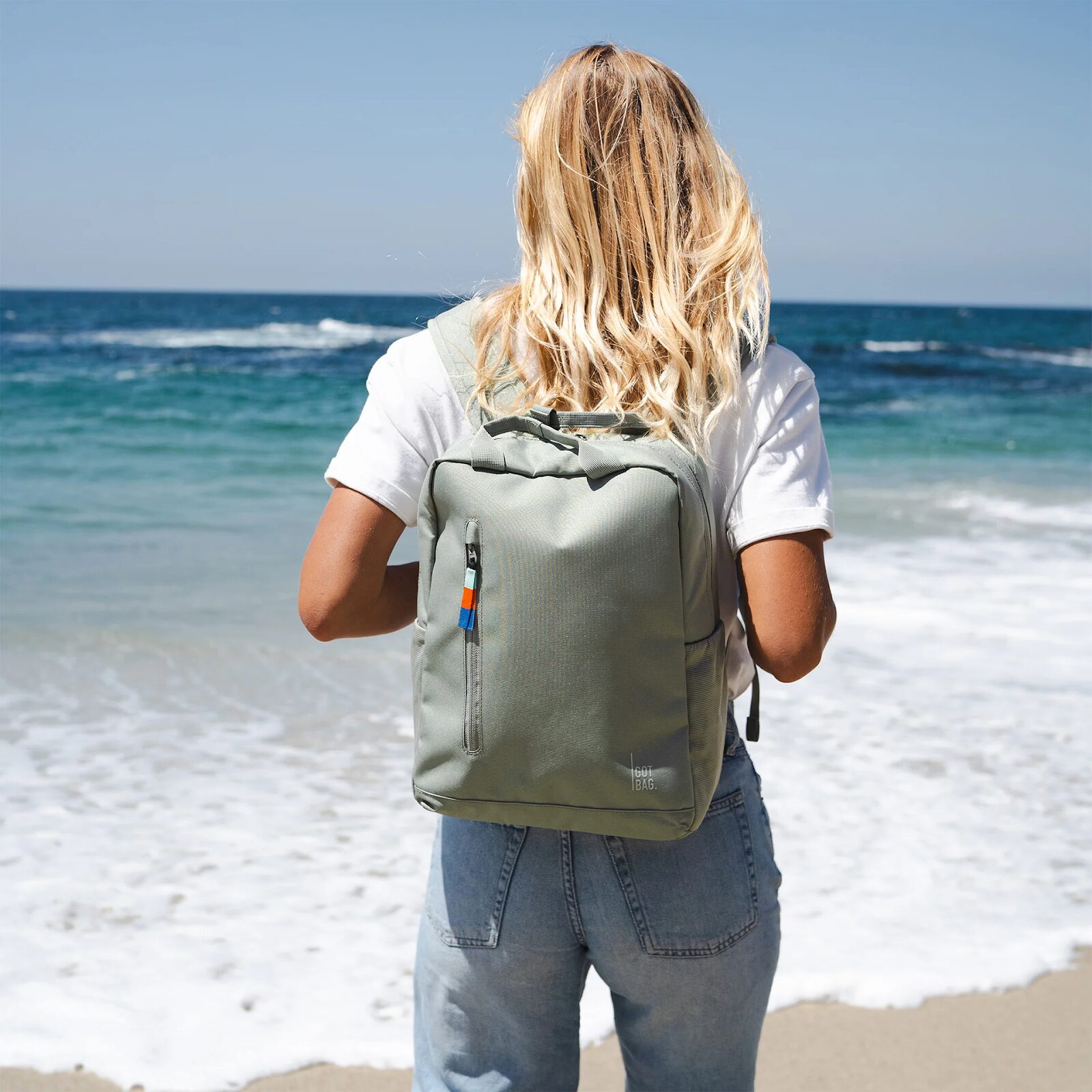 A blonde person wearing a minimalist backpack standing on a beach facing the ocean.