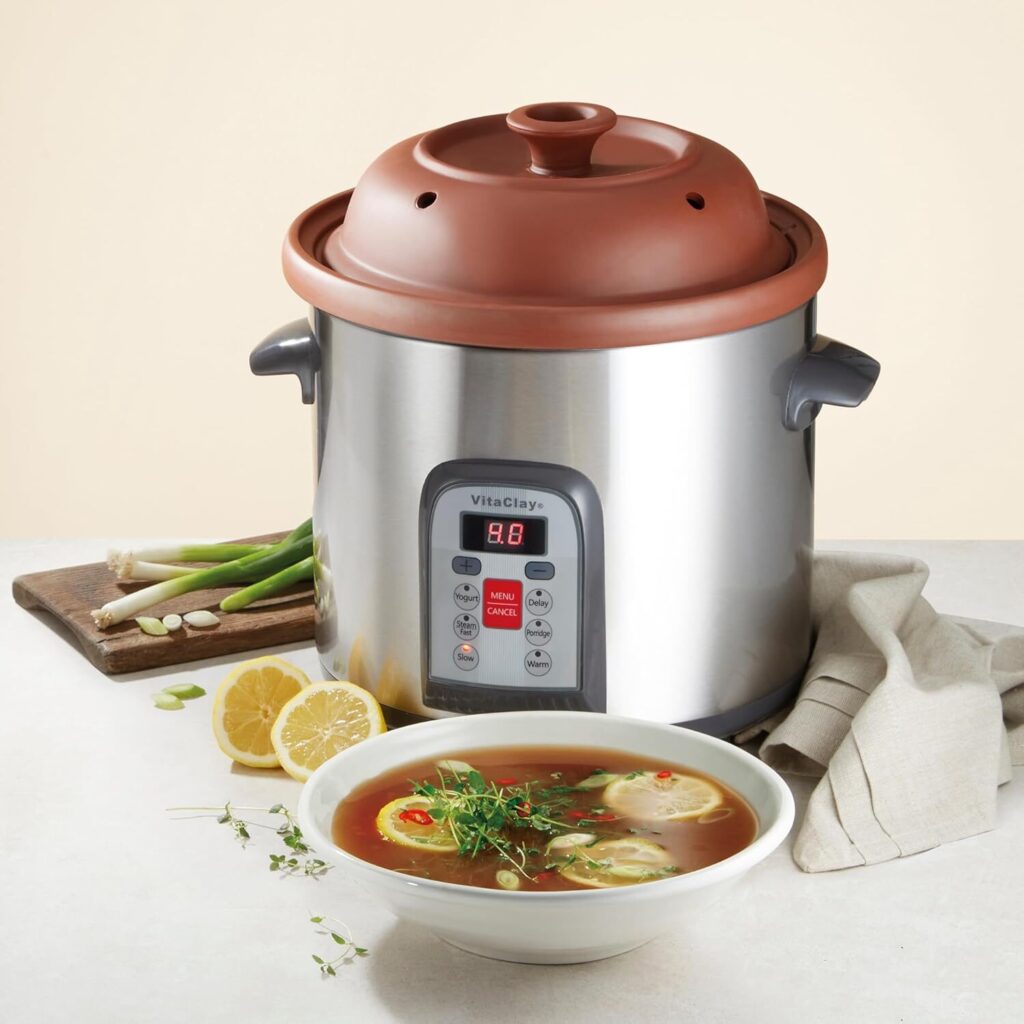 A Vitaclay crockpot on a counter with a cutting board, kitchen towel, and bowl of soup.