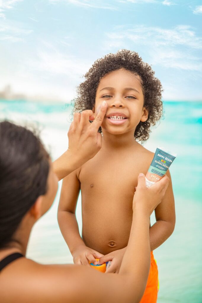 A parent rubbing Happy Ocean brand sunscreen on a child’s face in front of the ocean.
